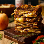 Stack of grilled cheese with a fork, apple, and hot honey in background.