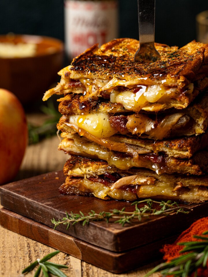 Stack of grilled cheese with a fork, apple, and hot honey in background.