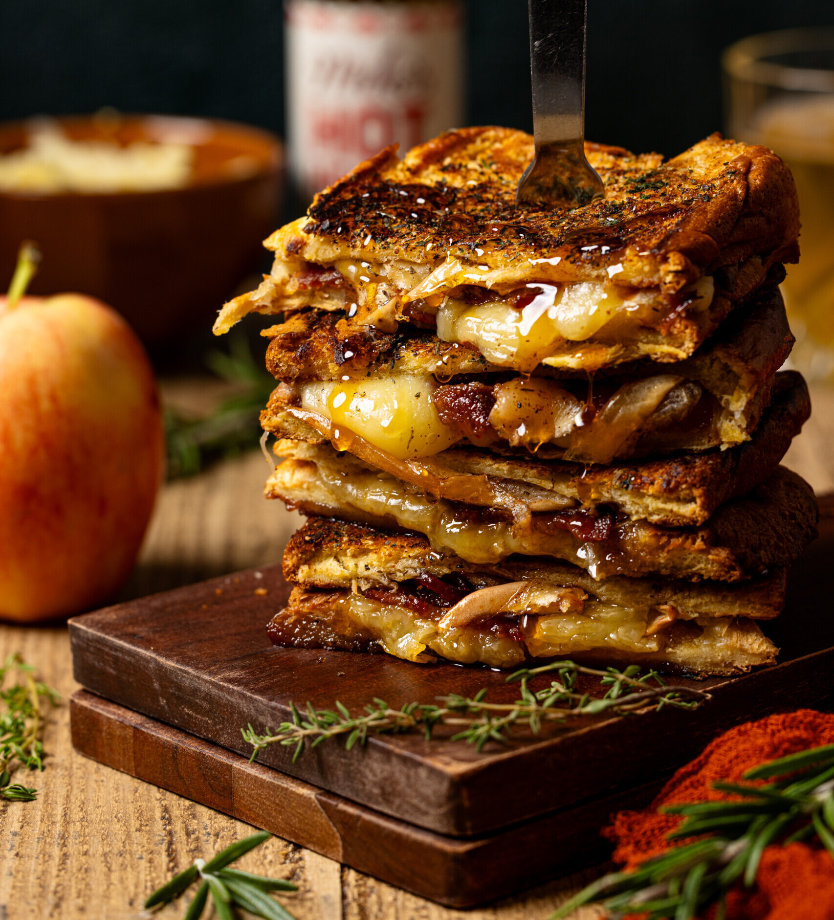 Stack of grilled cheese with a fork, apple, and hot honey in background.