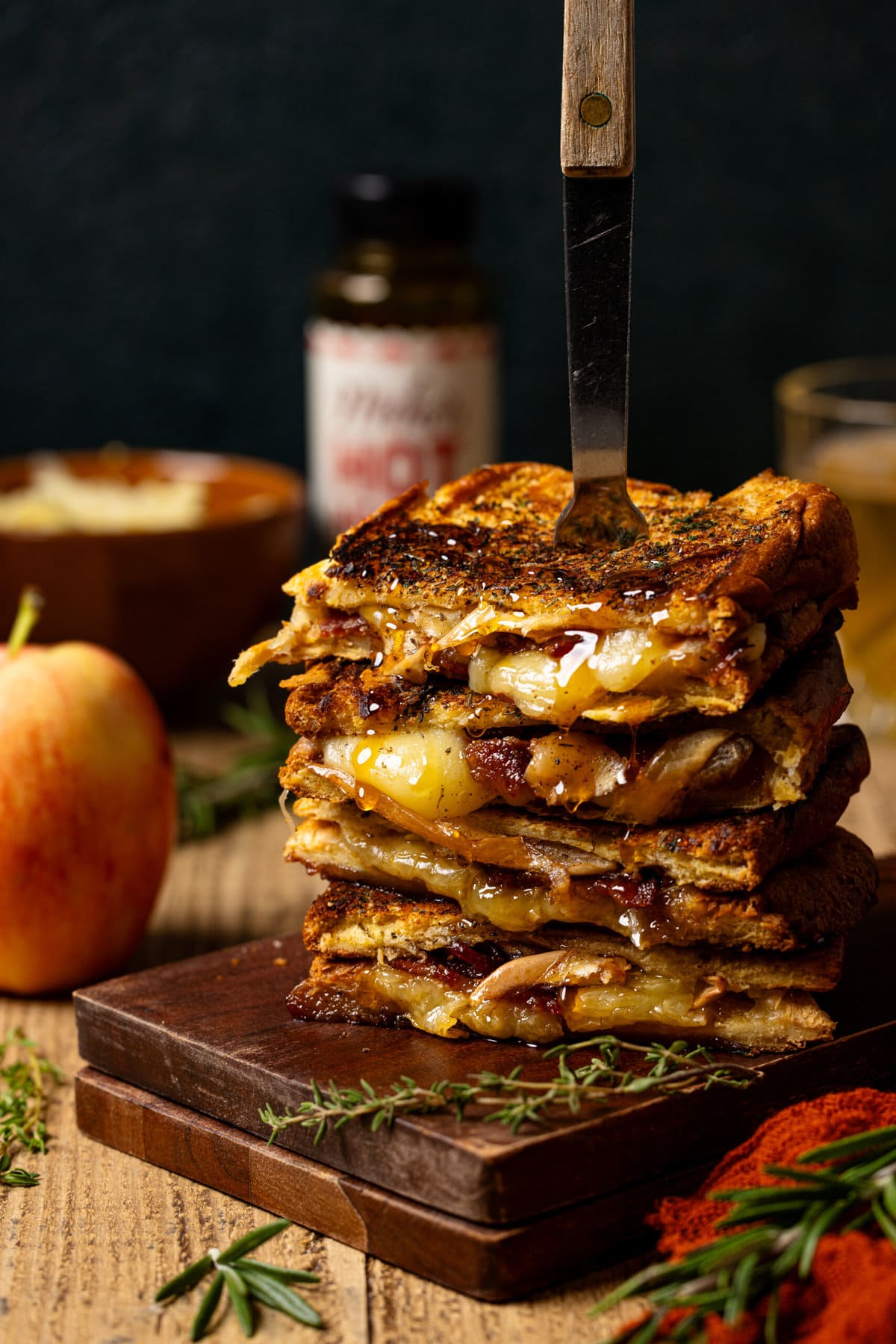 Stack of grilled cheese with a fork, apple, and hot honey in background.