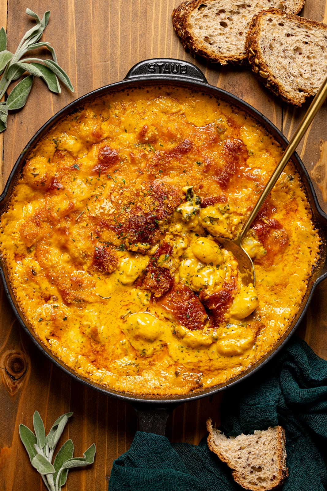 Baked gnocchi in a black skillet with a gold spoon, herbs, and pieces of bread. 