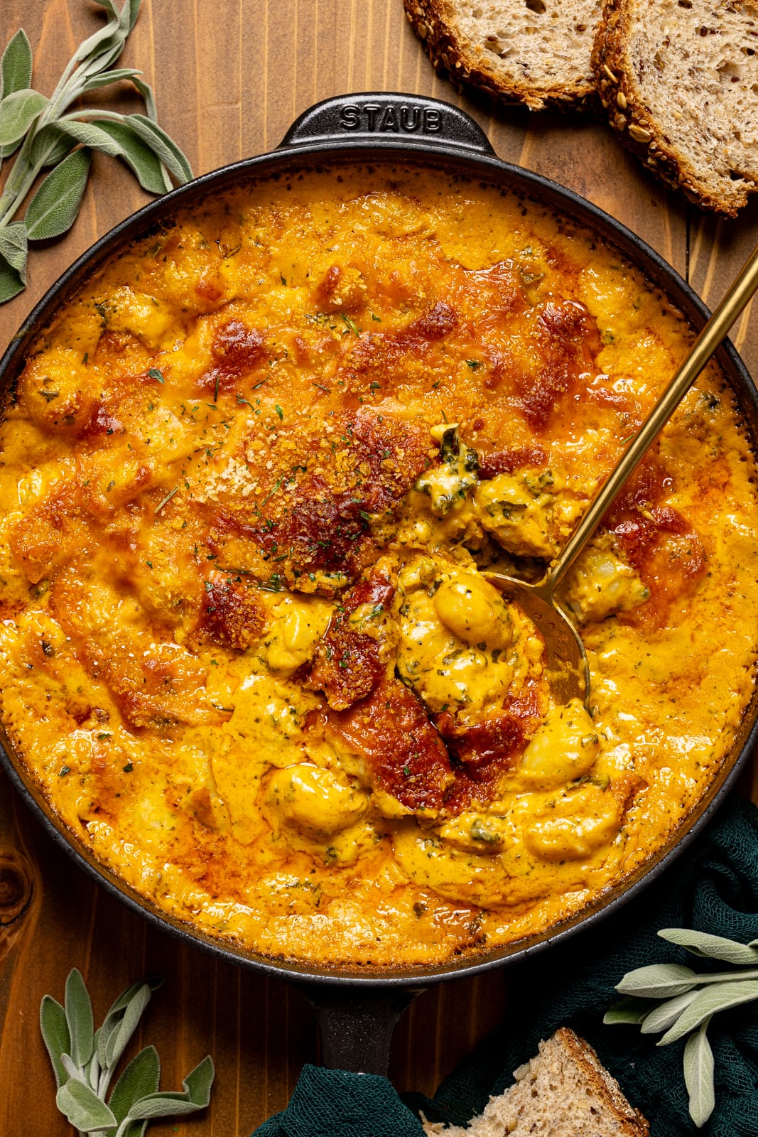 Baked gnocchi in a black skillet with a gold spoon, herbs, and pieces of bread.