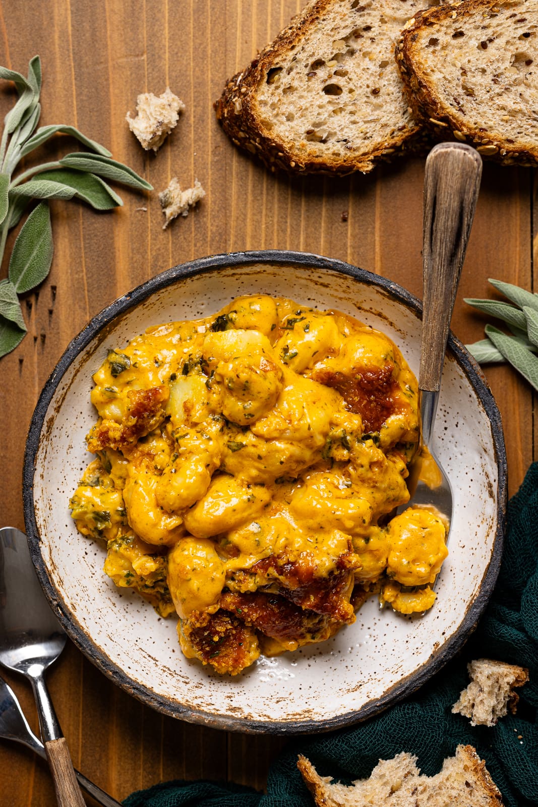 Baked gnocchi in a white bowl with a fork and spoon on a brown wood table with bread and herbs. 