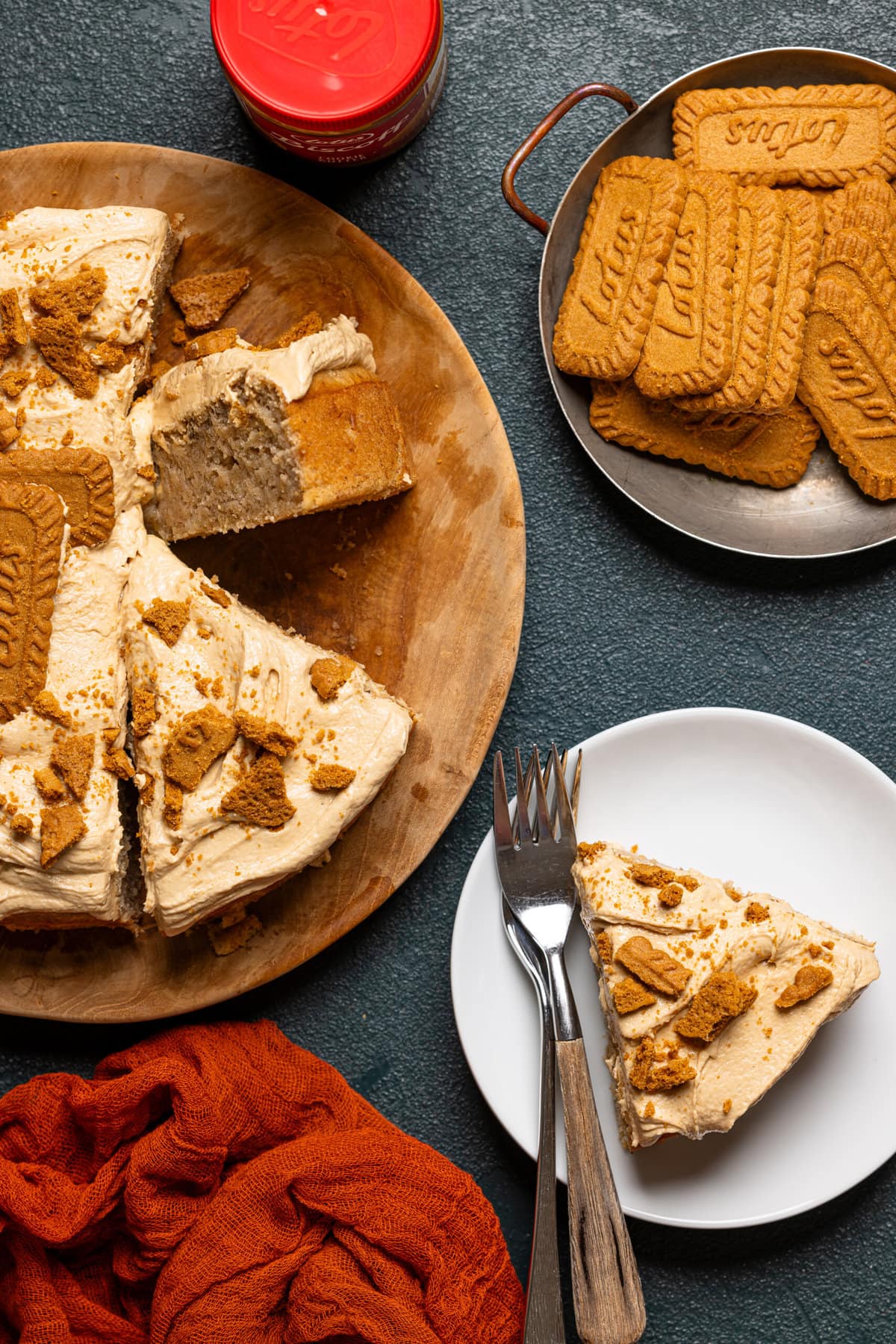 Slice of banana cake on a white plate with two forks with whole cake on a wooden cake platter.