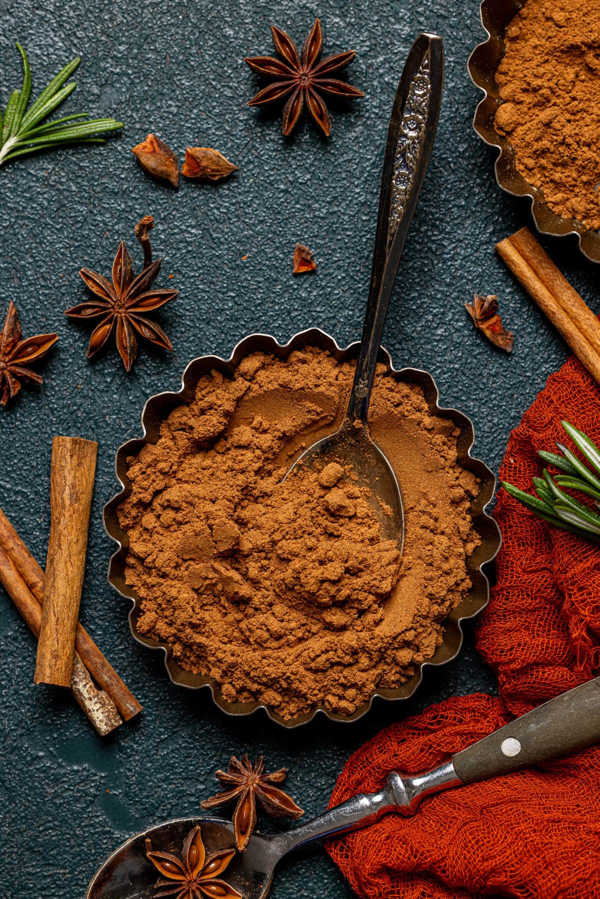 Pumpkin spice blend in a small tin with a spoon, cinnamon stick, and spices as garnish.