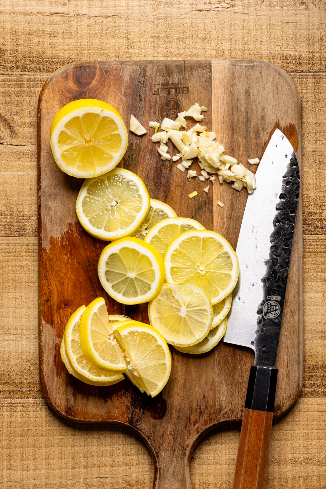 Lemon slices and minced garlic on a wood cutting board with a knife.