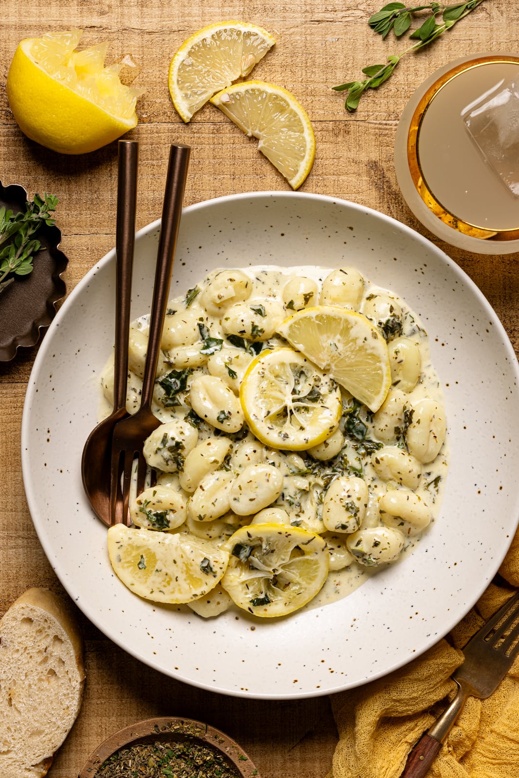 Gnocchi in a plate with a fork + spoon with a side of drink, lemons, and herbs.