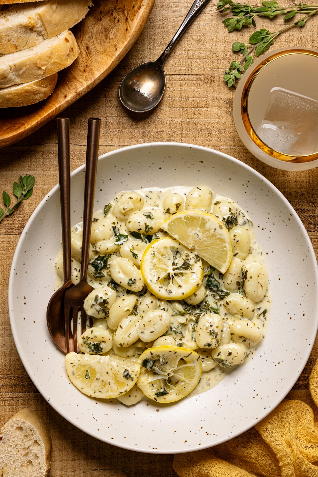 The best herb butter gnocchi in a white plate with fork + spoon, side of bread, drink, and lemons.