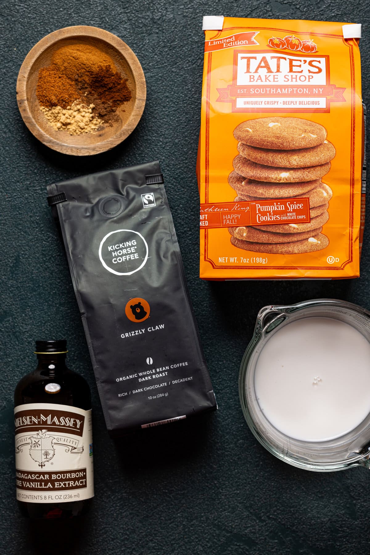 Ingredients on a dark green table including cookies, spices, coffee beans, vanilla, and milk.