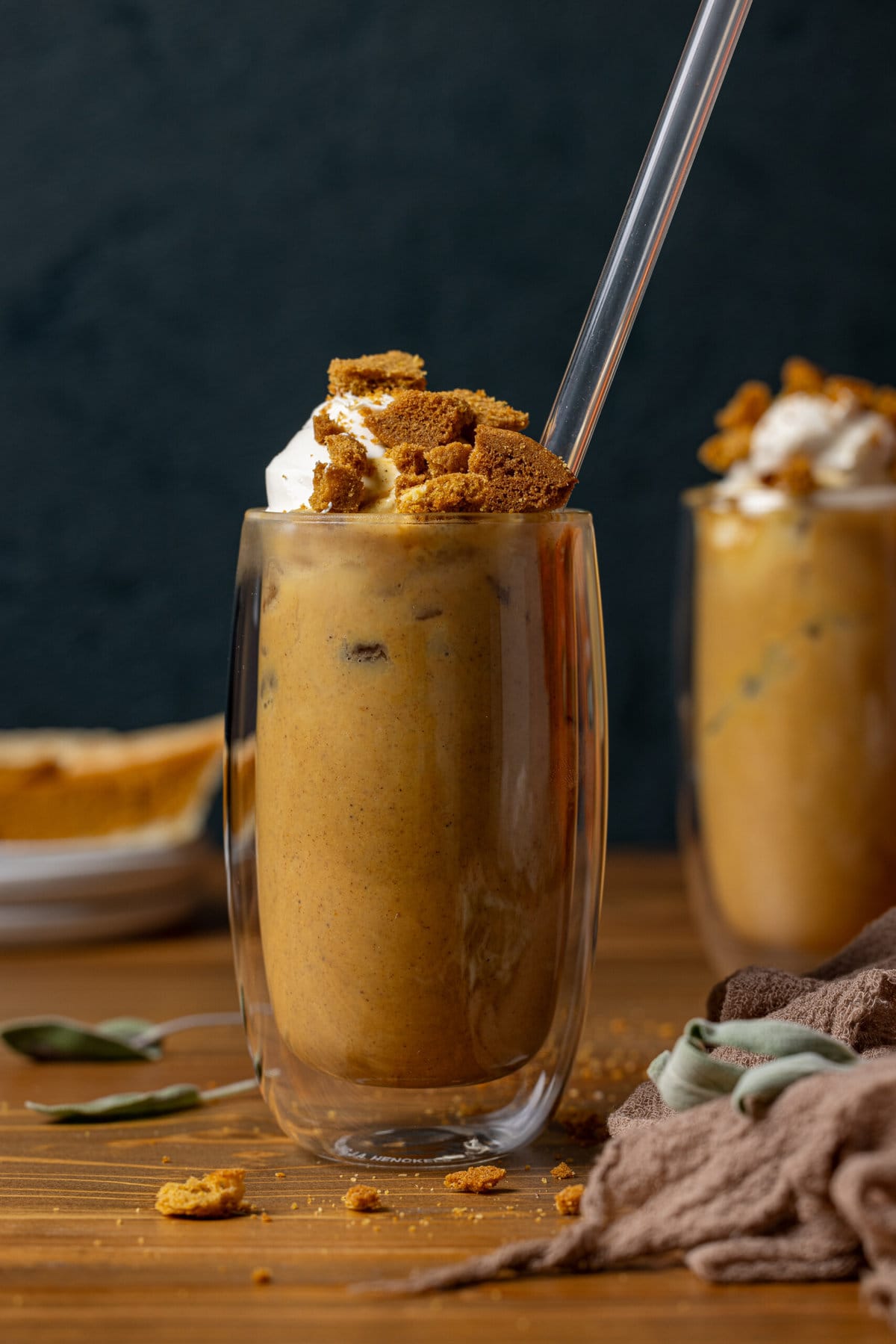 Latte drinks in clear glasses with a straw and slice of pumpkin pie in the background. 