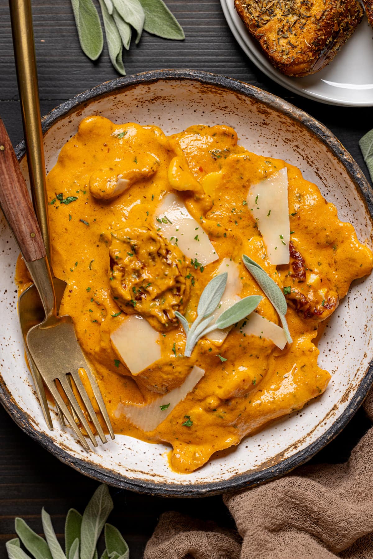 Ravioli in a bowl with two forks and a garnish of parmesan shavings and sage leaves.