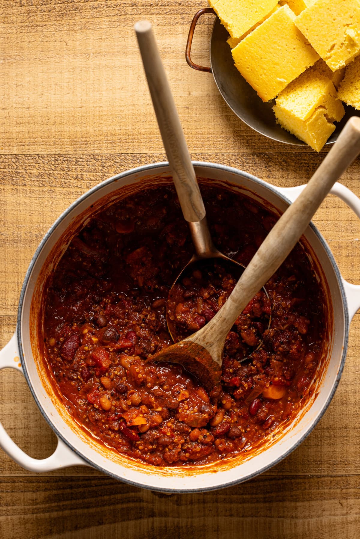 Pot of chili with two serving spoons and side of cornbread. 