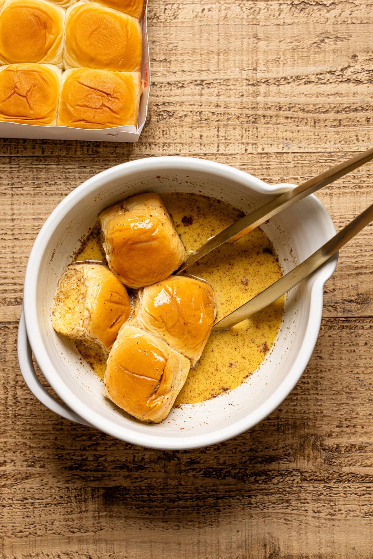 Hawaiian Rolls soaked in the batter in a white bowl with tongs.