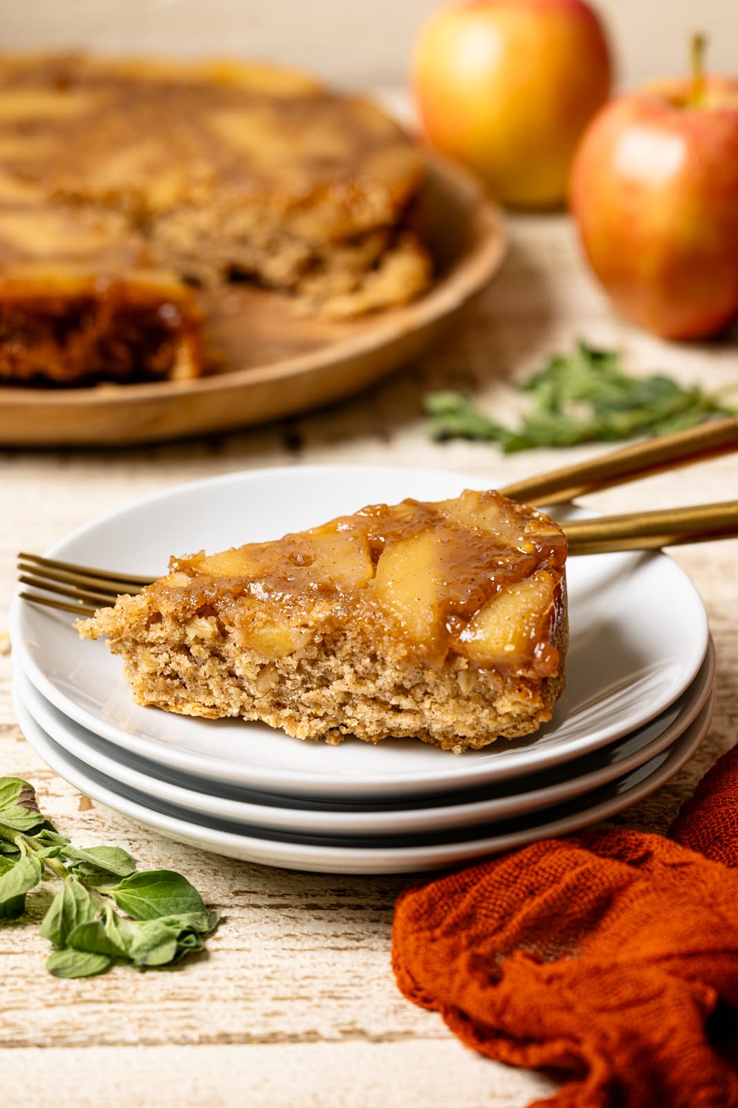 Slice of cake on stacked white plates with apples in the background. 