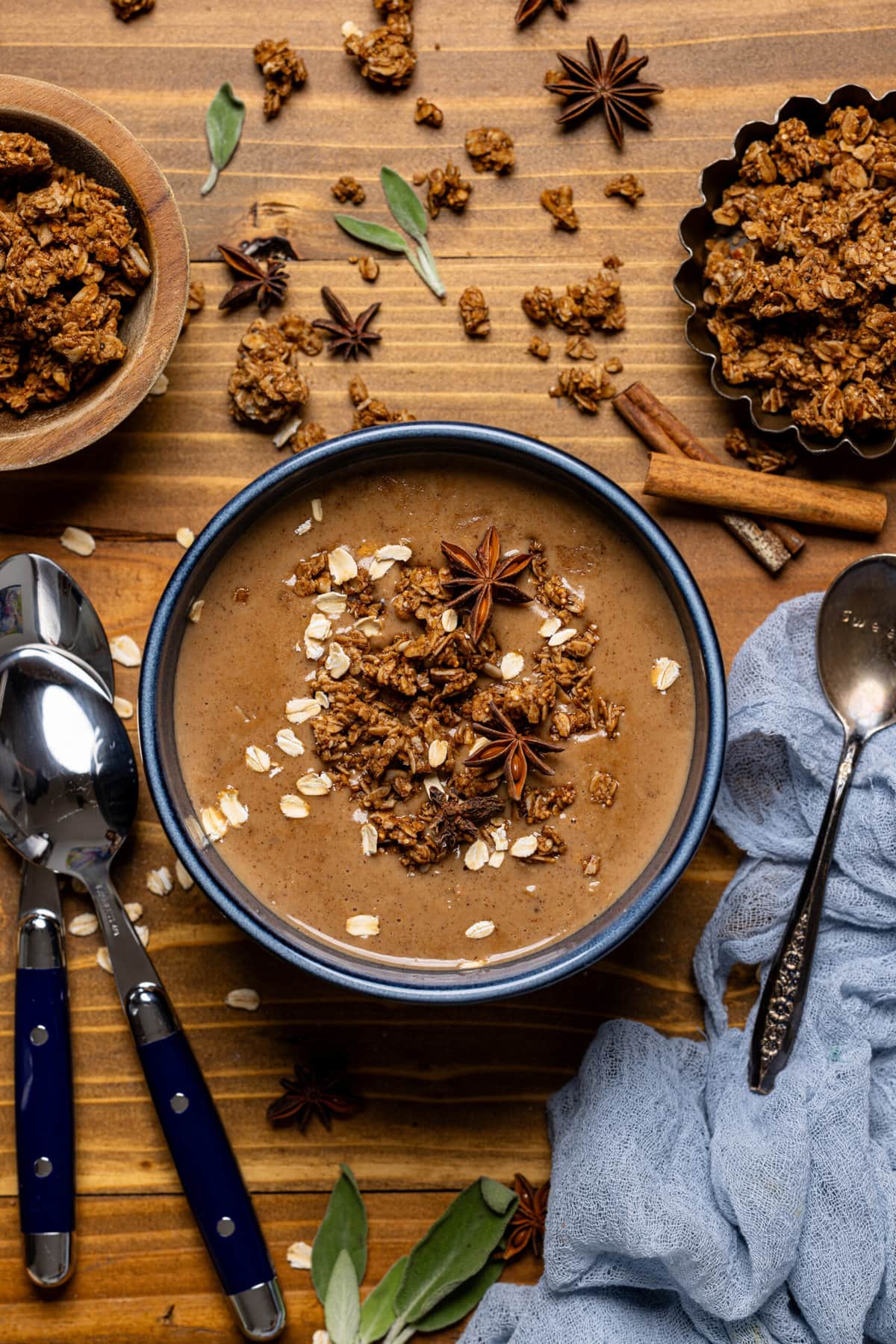 Bowl of dirty chai latte oatmeal with spoons, granola, and a blue napkin on a wood table.