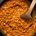 Cooked pinto beans in BBQ sauce in a deep black skillet and wooden spoon.