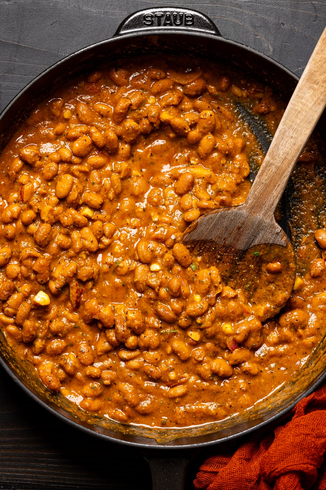 Cooked pinto beans in BBQ sauce in a deep black skillet and wooden spoon.