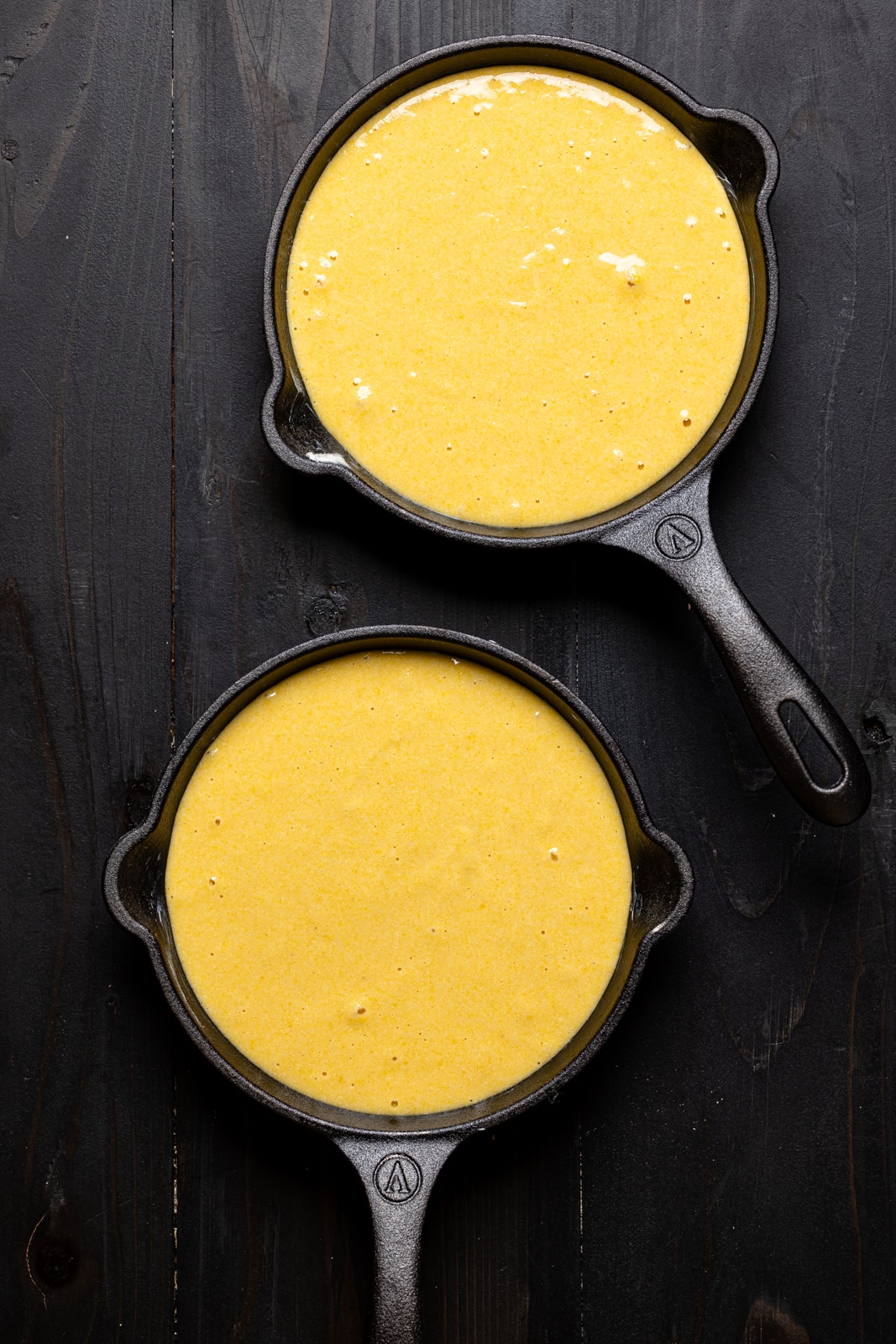 Hot honey cornbread batter in two black mini skillets on a black wood table.