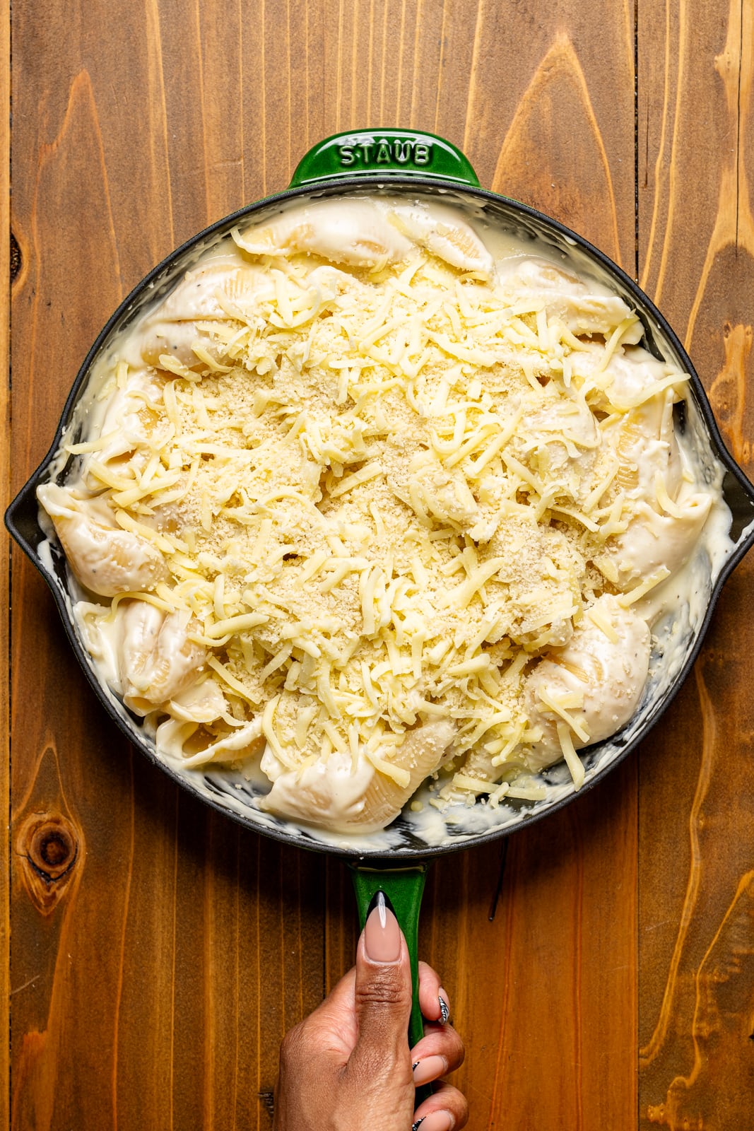 Assembled mac and cheese in a green skillet being held on a brown wood table.