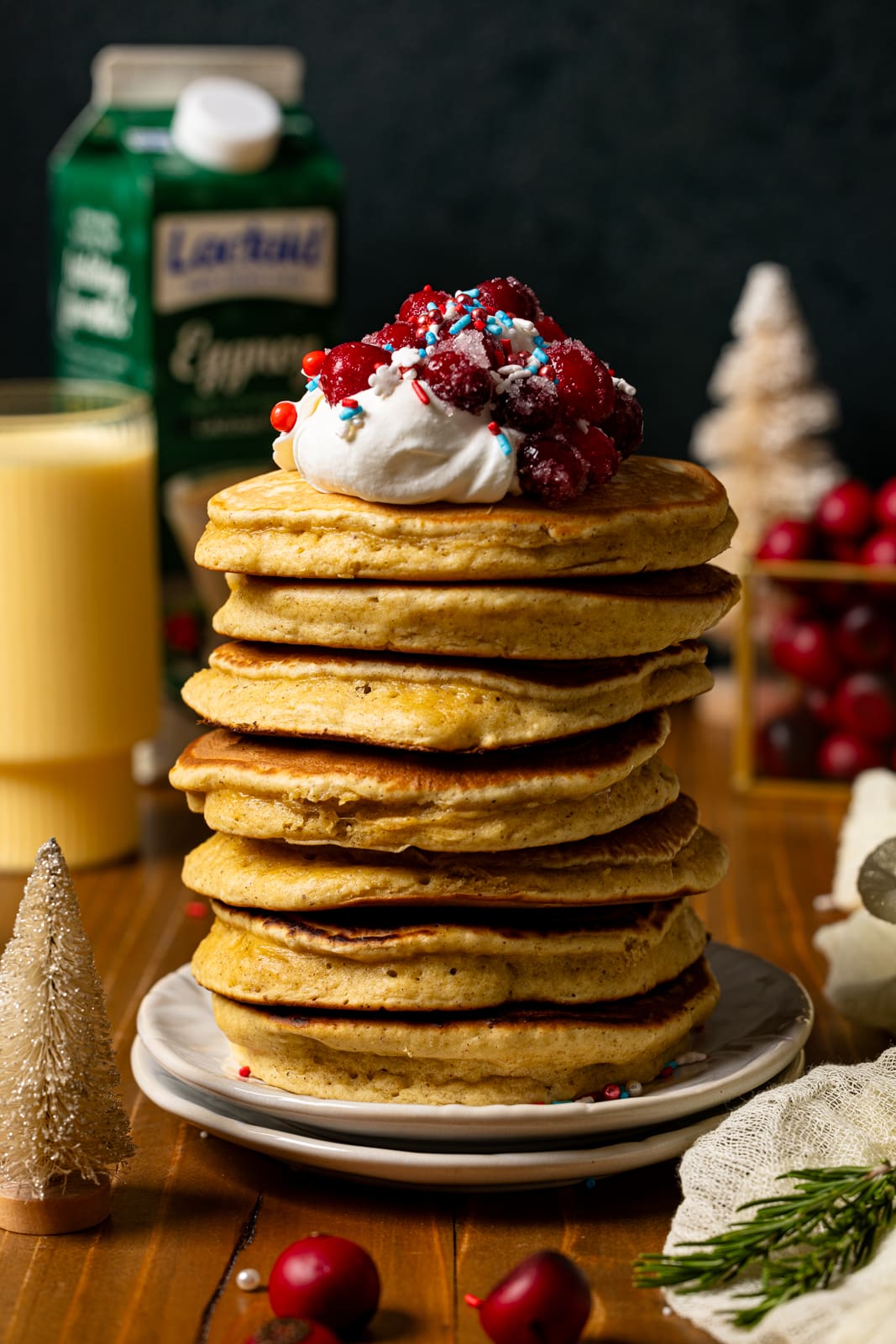 Stack of pancakes with eggnog and holiday decor in background.