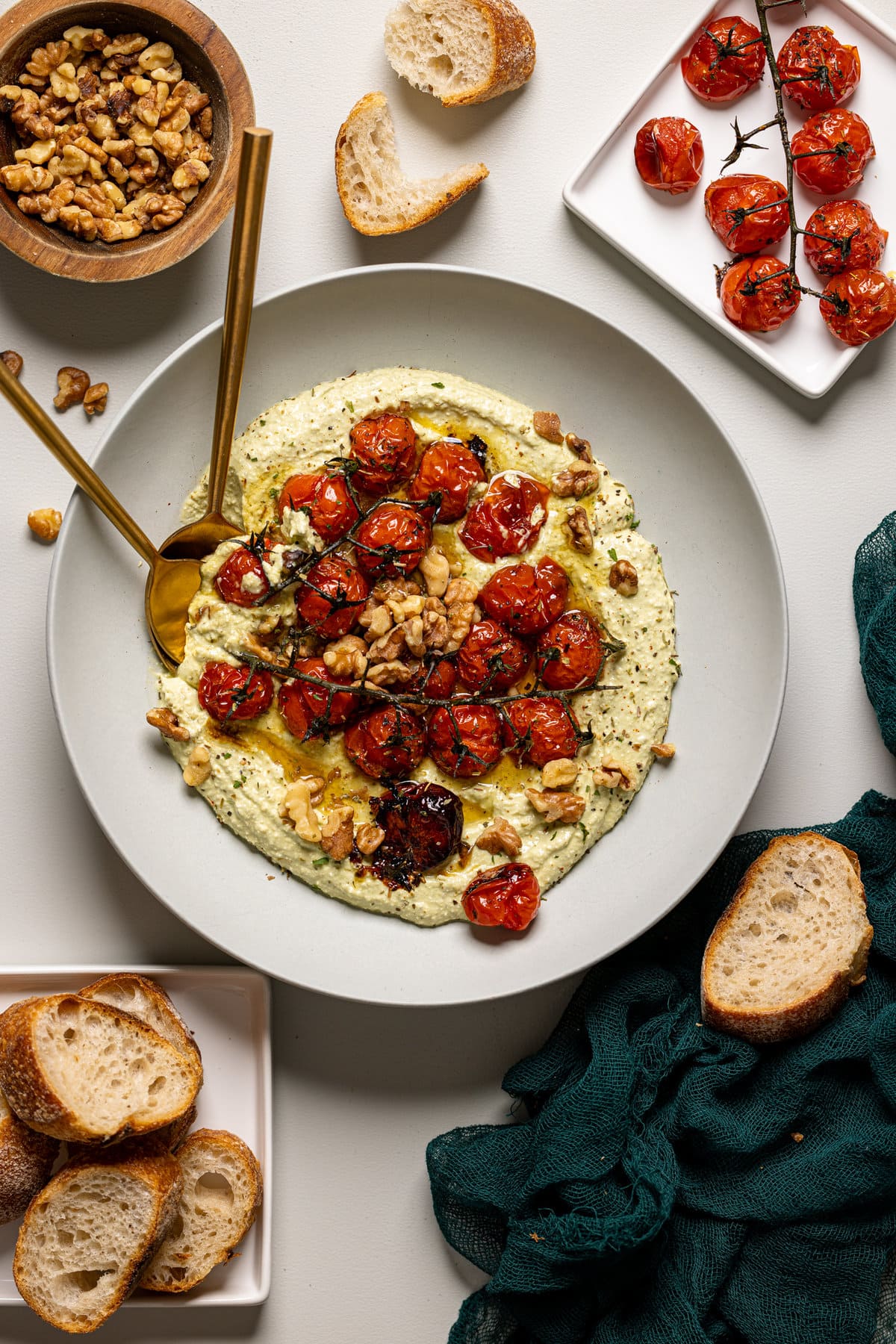 Overview shot of feta dip in a white low bowl with a spoon, fork, side of roasted tomatoes, bread, and nuts.