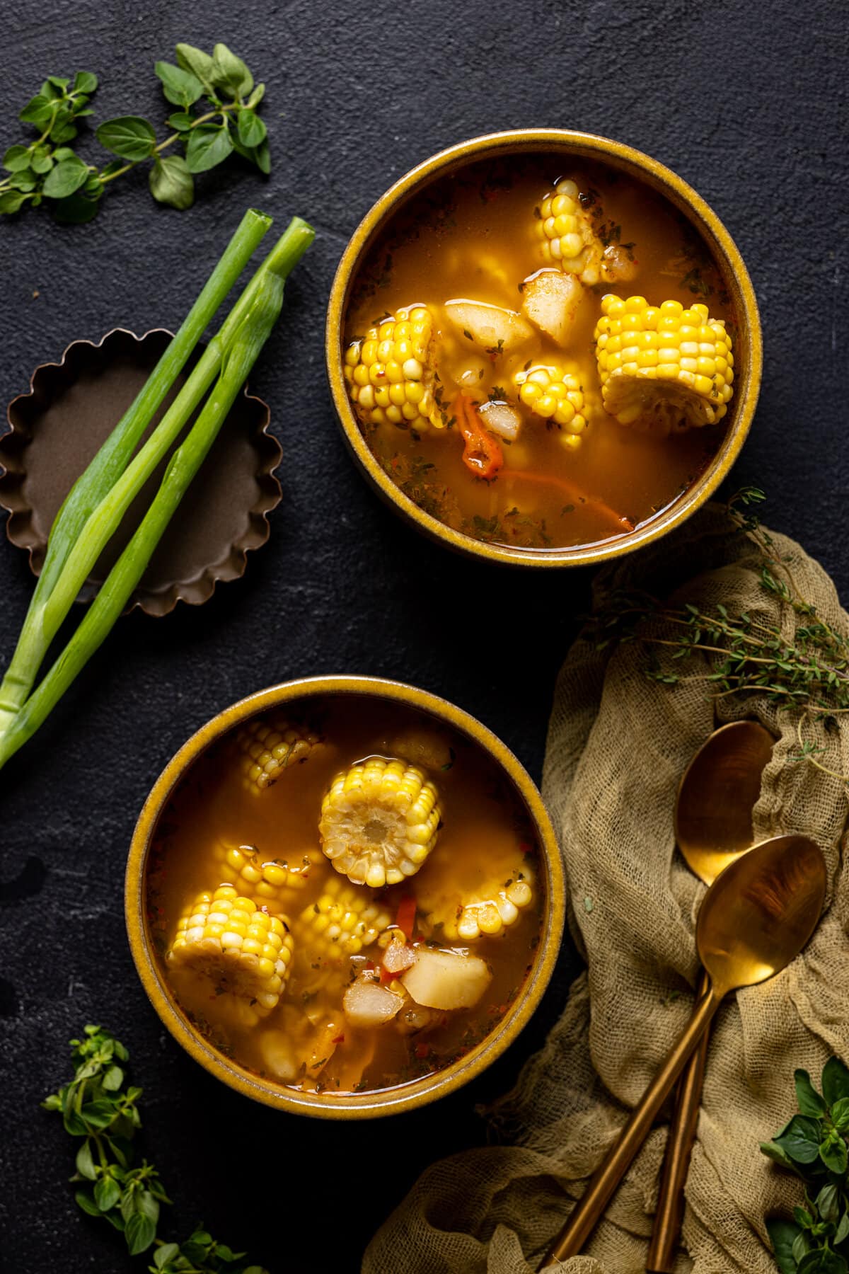 Two bowls of soup on a black table with a spoon and veggies. 