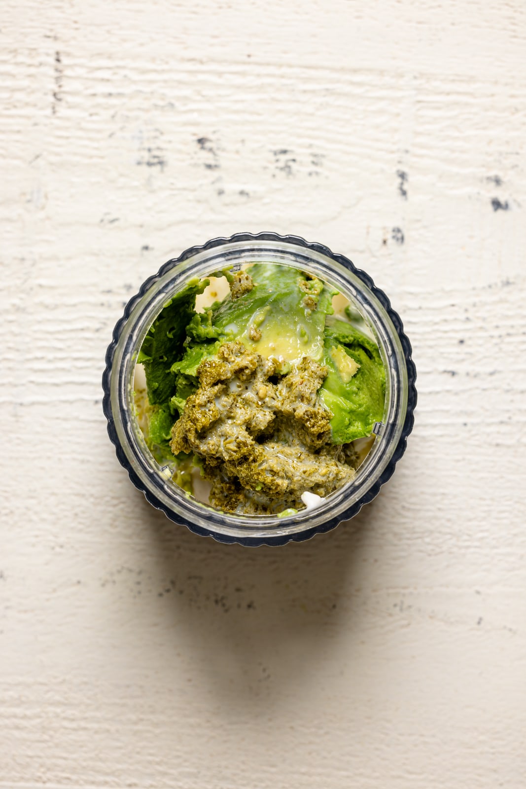 All ingredients for pesto avocado mayo in a blender on a white wood table.