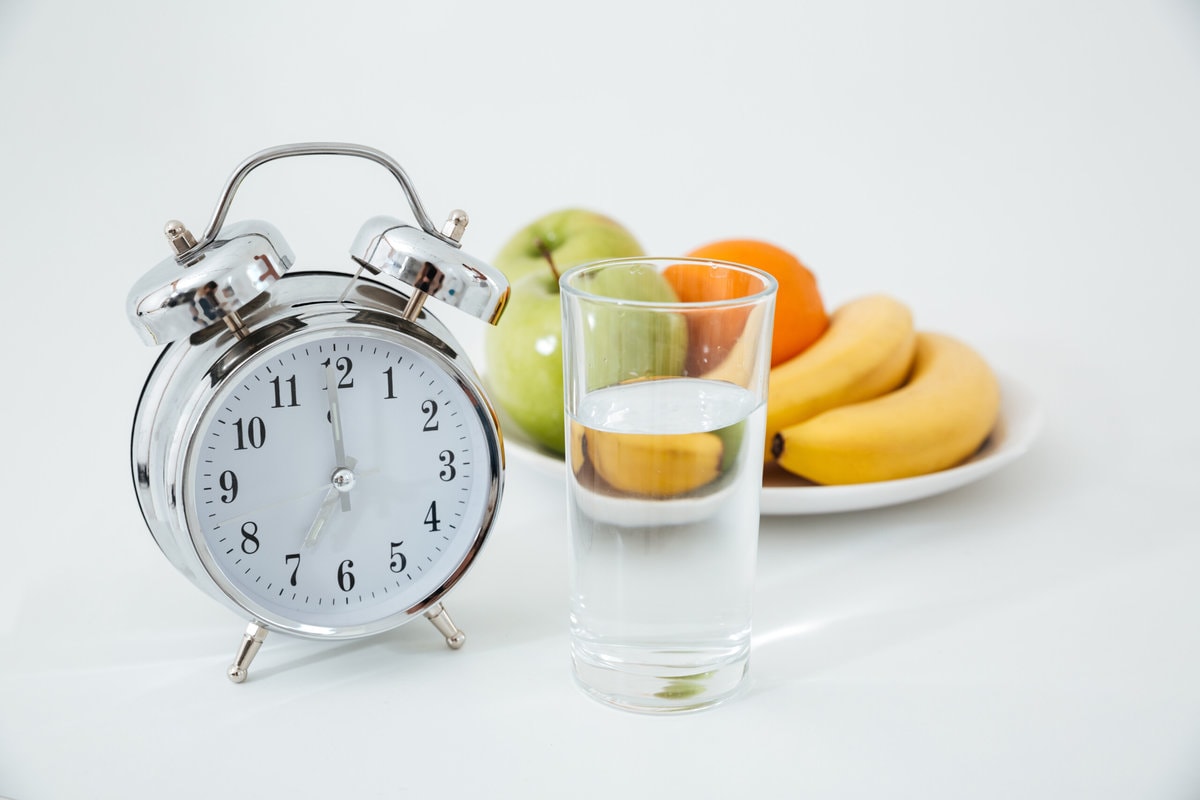 An Alarm Clock Next To A Glass Of Water And Bowl Of Fruit