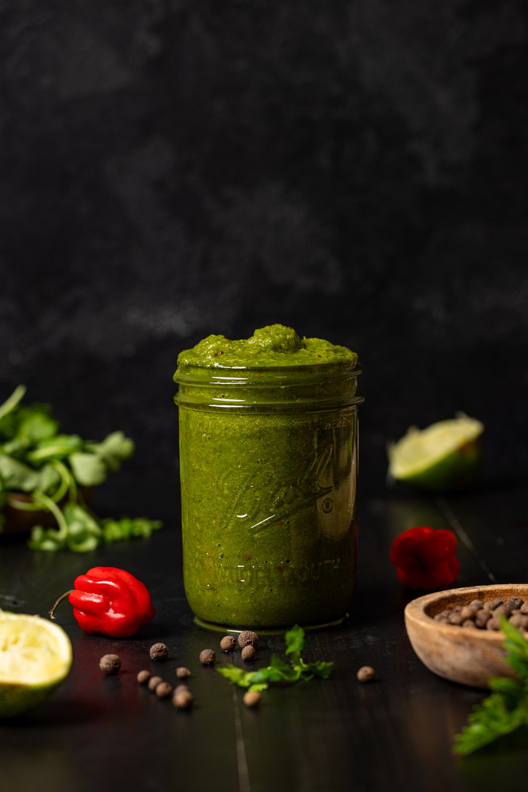 Green seasoning in a mason jar with vegetables surrounding on a black wood table.