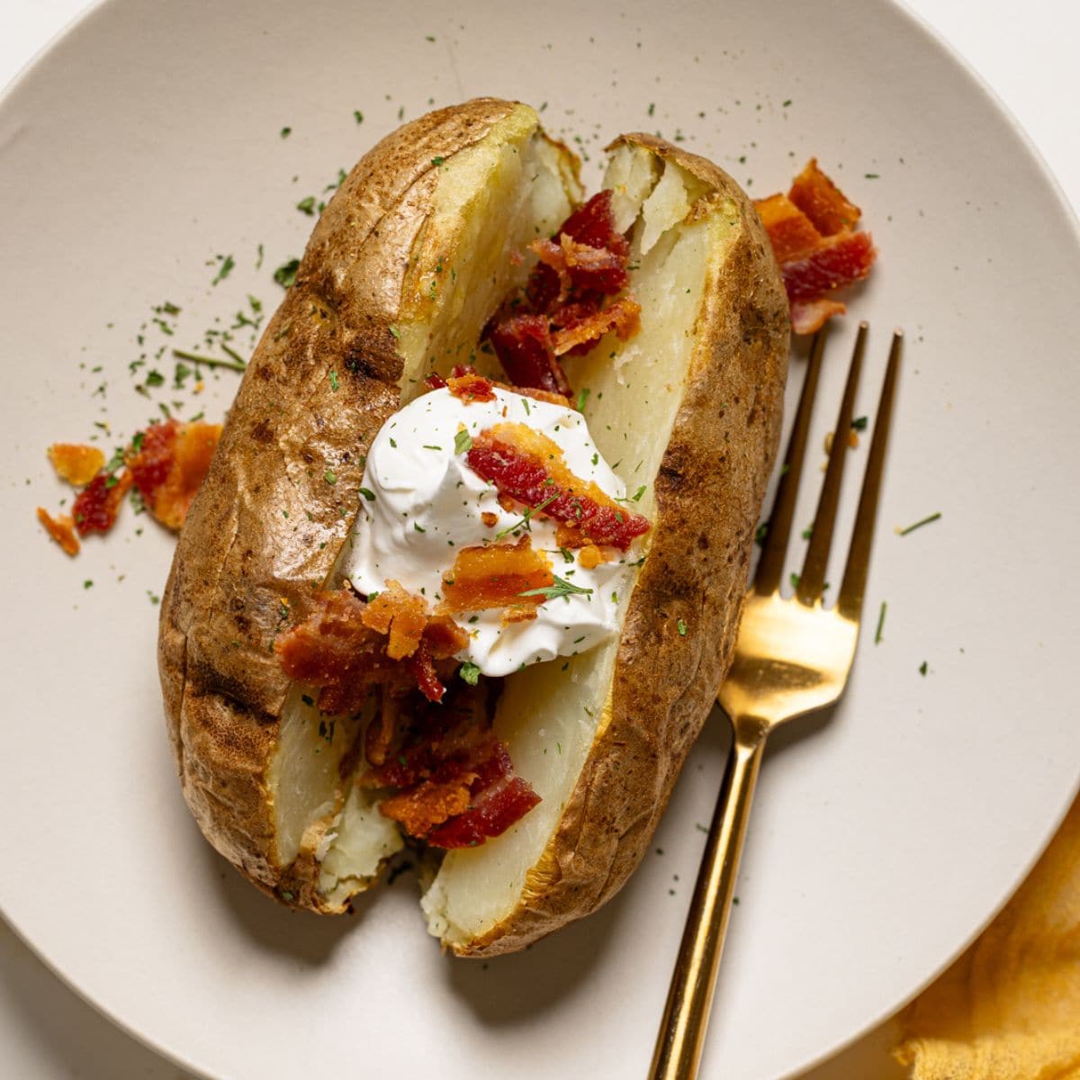 Baked potato on a white plate with a fork topped with bacon and sour cream.