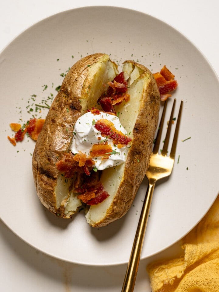 Baked potato on a white plate with a fork topped with bacon and sour cream.