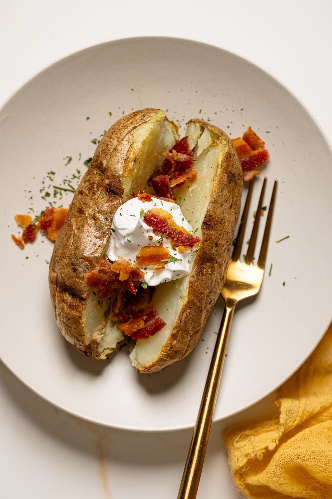 Baked potato on a white plate with a fork topped with bacon and sour cream.