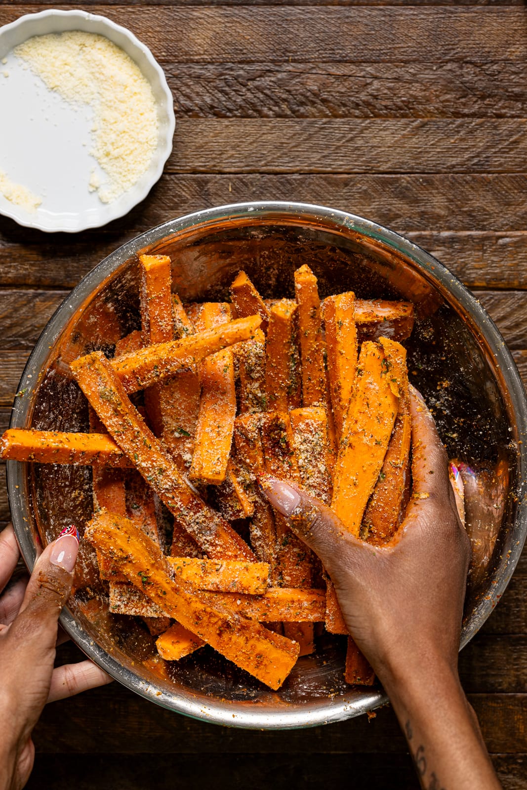 Fries in a bowl being seasoned with hands.