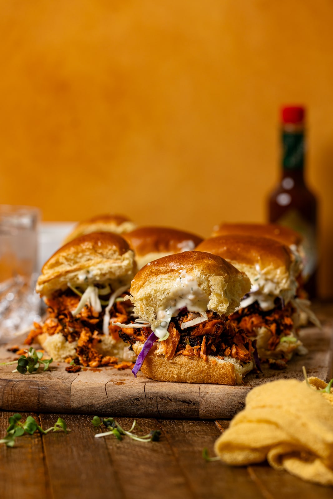 Sliders on a brown board with hot sauce, a drink, and yellow napkin.