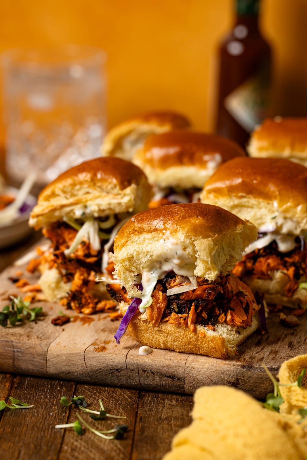 Sliders on a brown wood board with hot sauce and a yellow napkin.