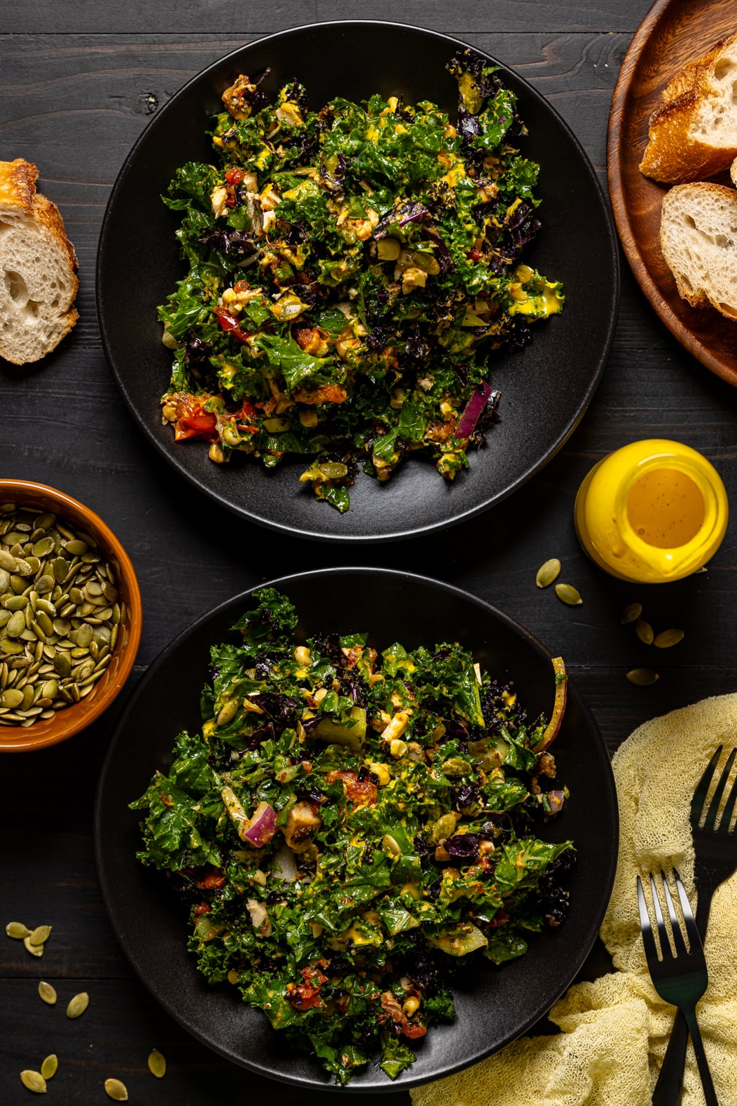 Salads on a two black plates with toppings, bread, and dressing on the side.