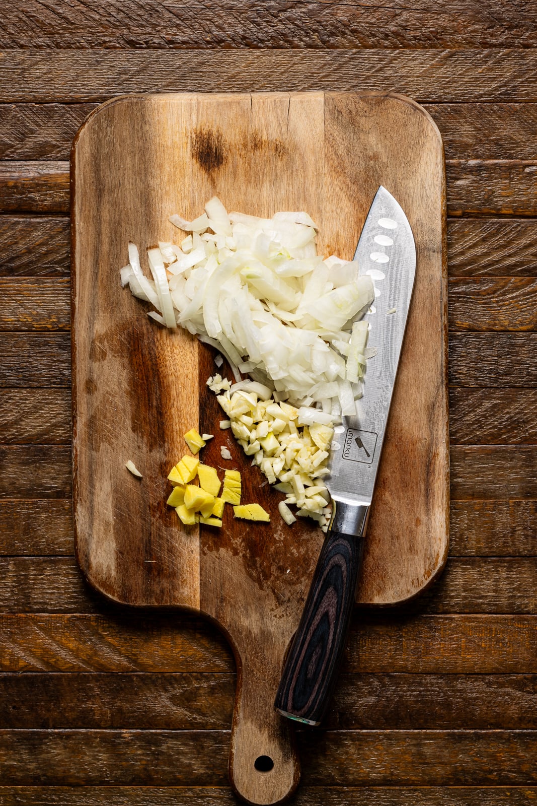 Chopped onions, garlic, and ginger on a cutting board with a knife.