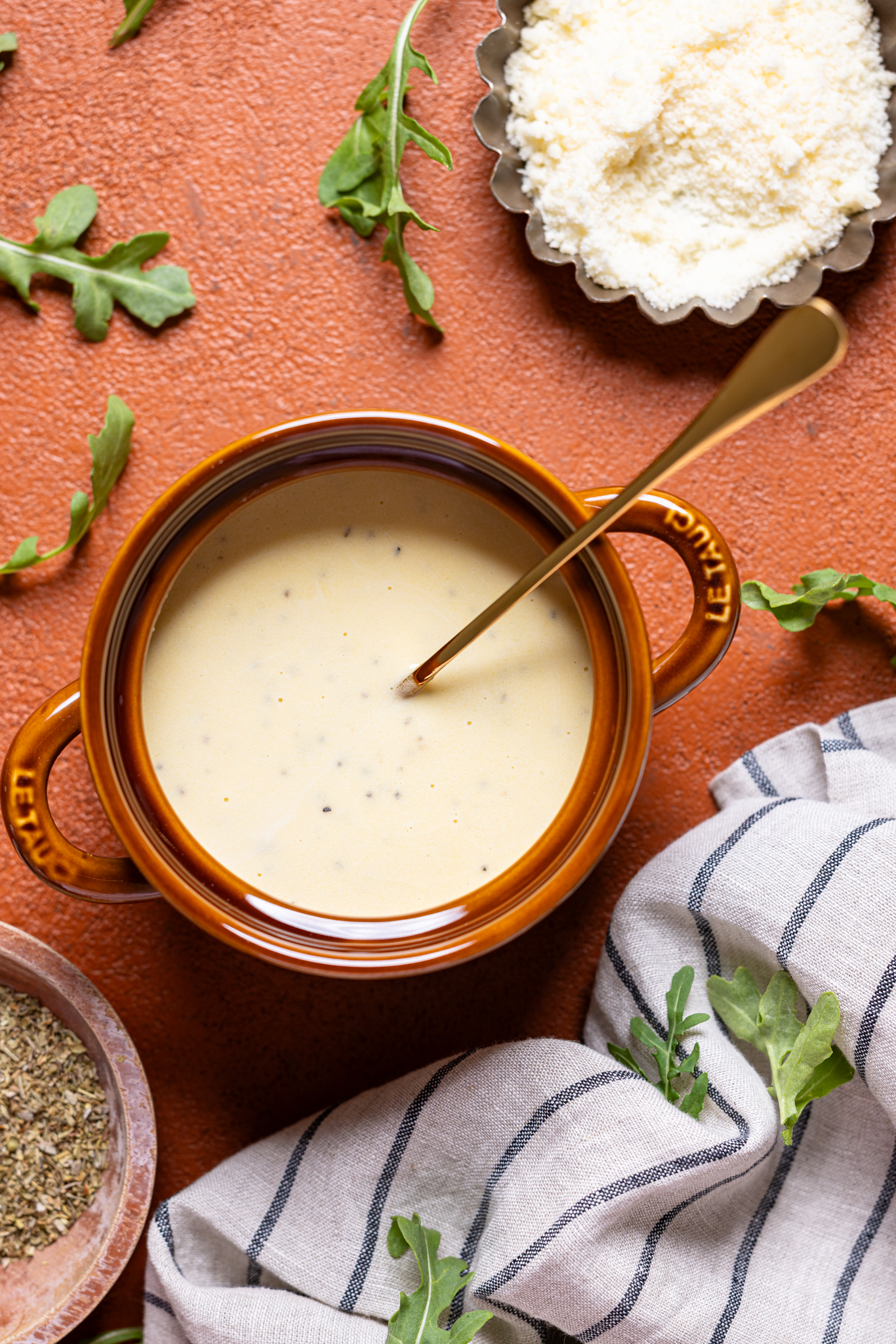Image of alfredo sauce in a brown bowl with a spoon and parmesan and herbs.