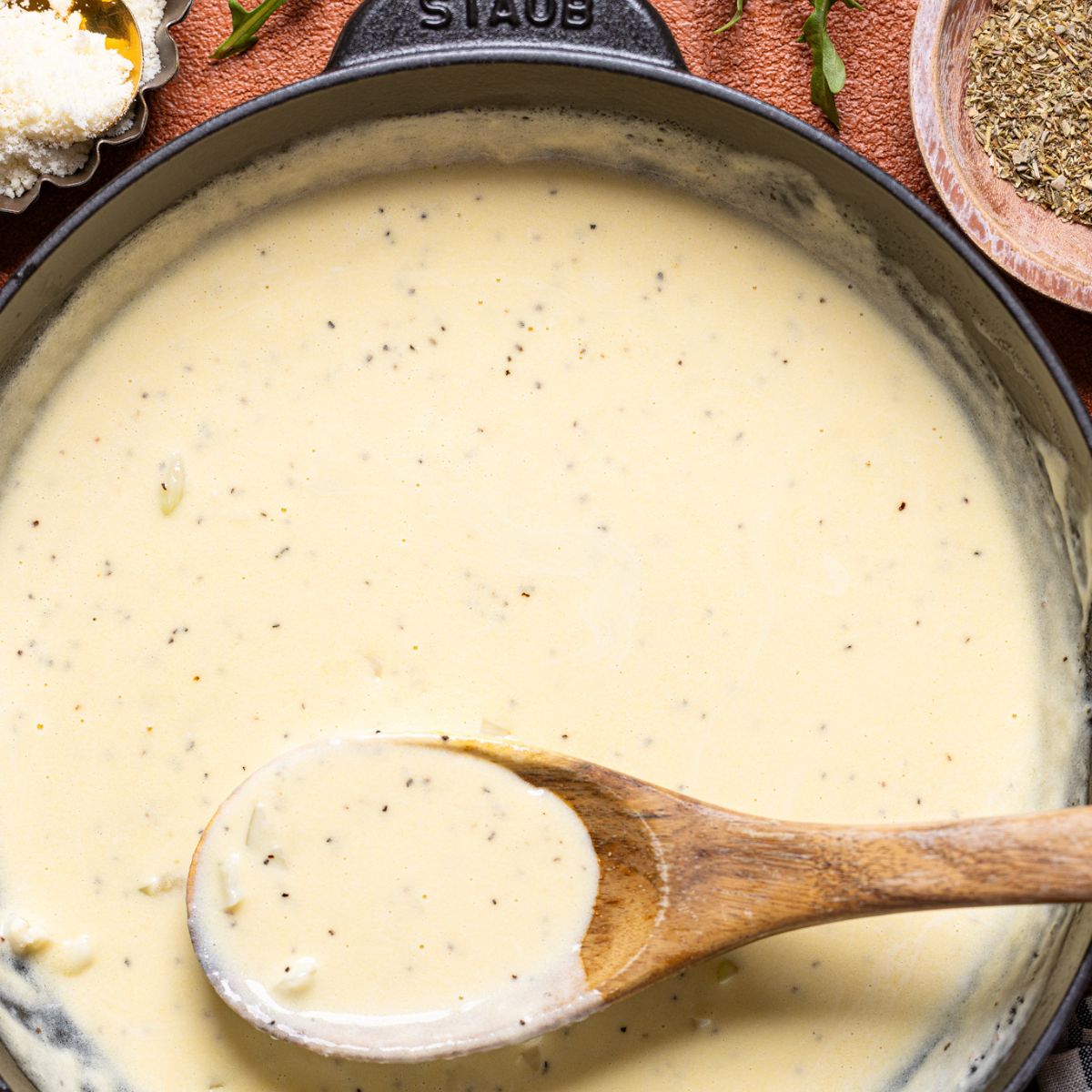 Image of alfredo sauce in a skillet with wooden spoon with parmesan.