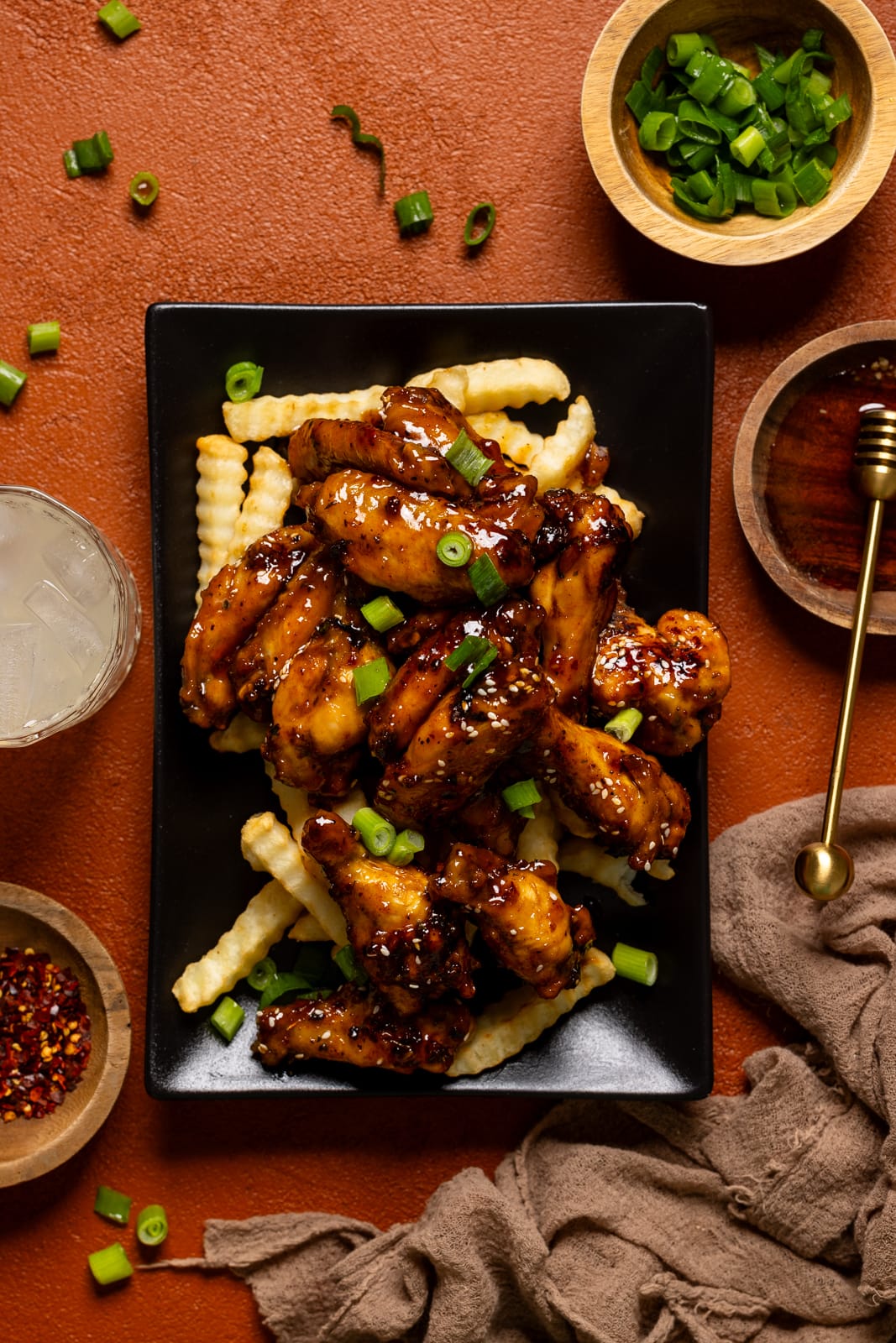 Chicken wings on a fries on a black platter with honey, scallions, and a drink on the side.
