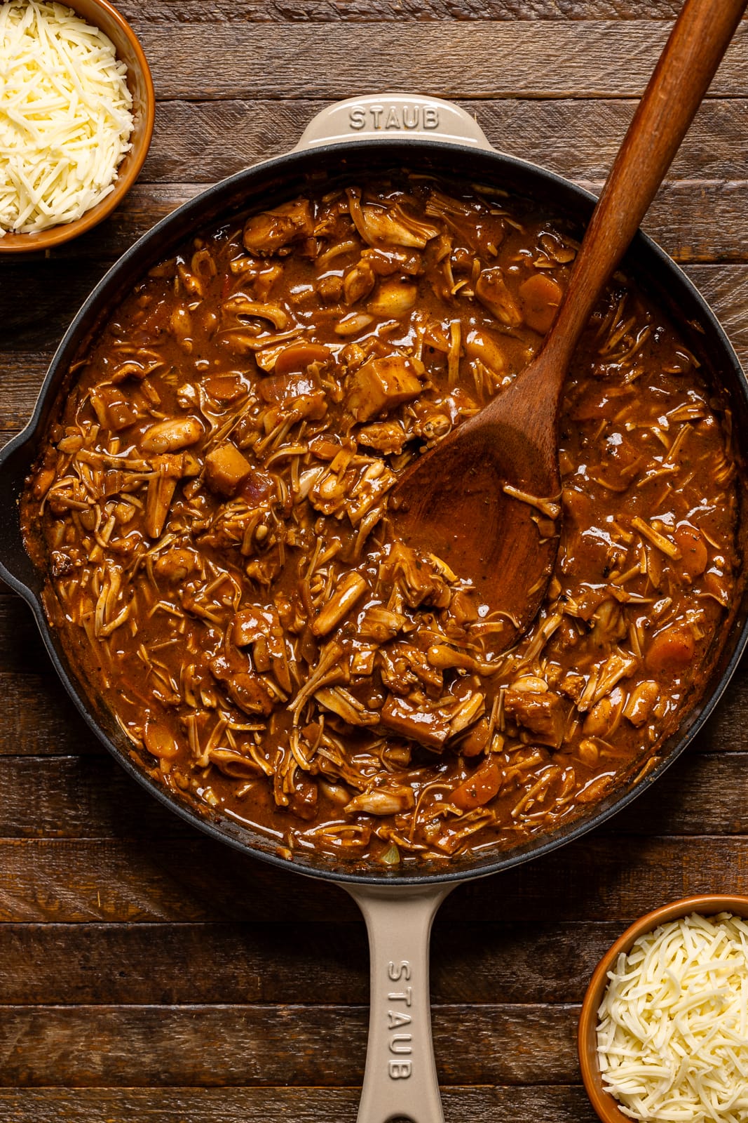 Cooked jackfruit in a beige skillet with a wooden spoon and bowls of cheese.