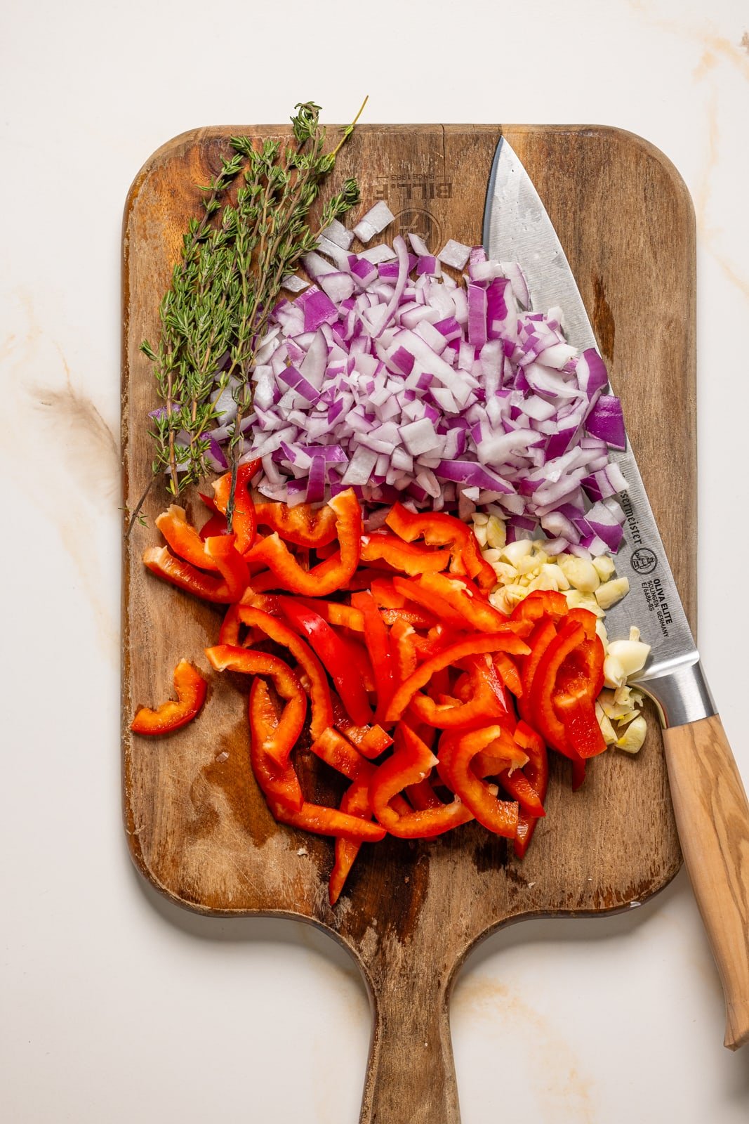 Bell peppers, onions, garlic, and thyme chopped on a cutting board with a knife.