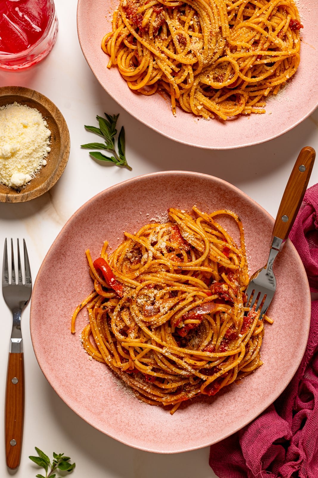 Spaghetti in two pink plates with a fork and a fork on the side and parmesan cheese.