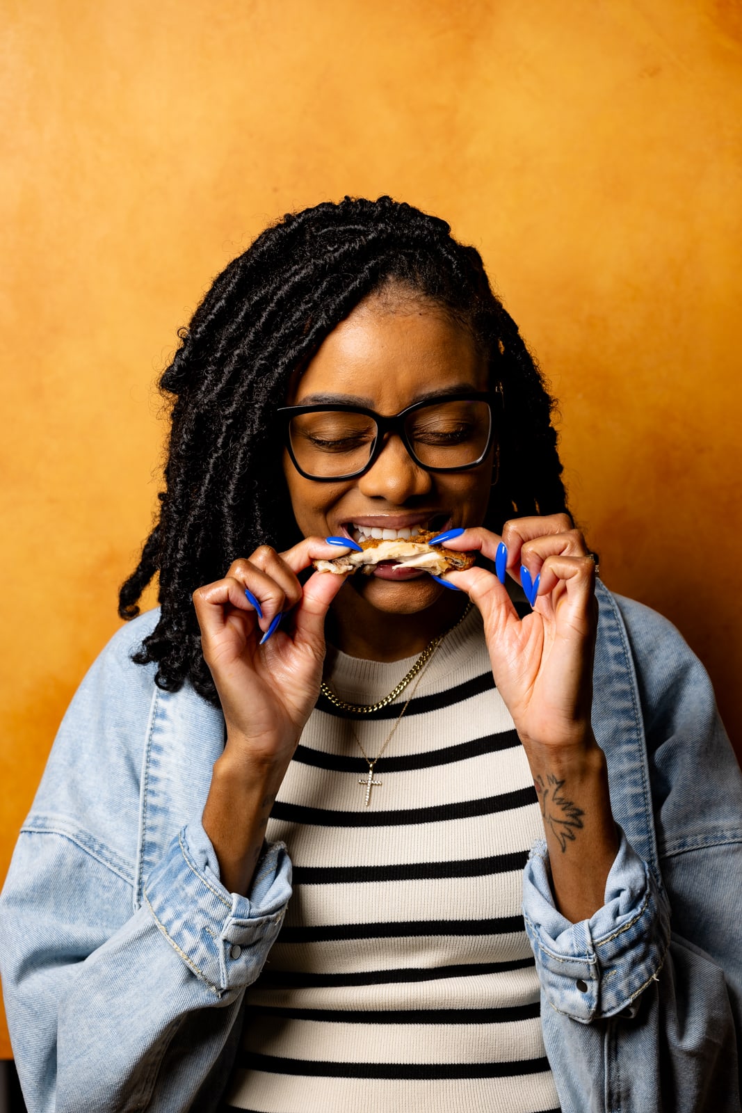 Picture of Shanika bitting a piece of chicken with her eyes closed in front of an orange background.