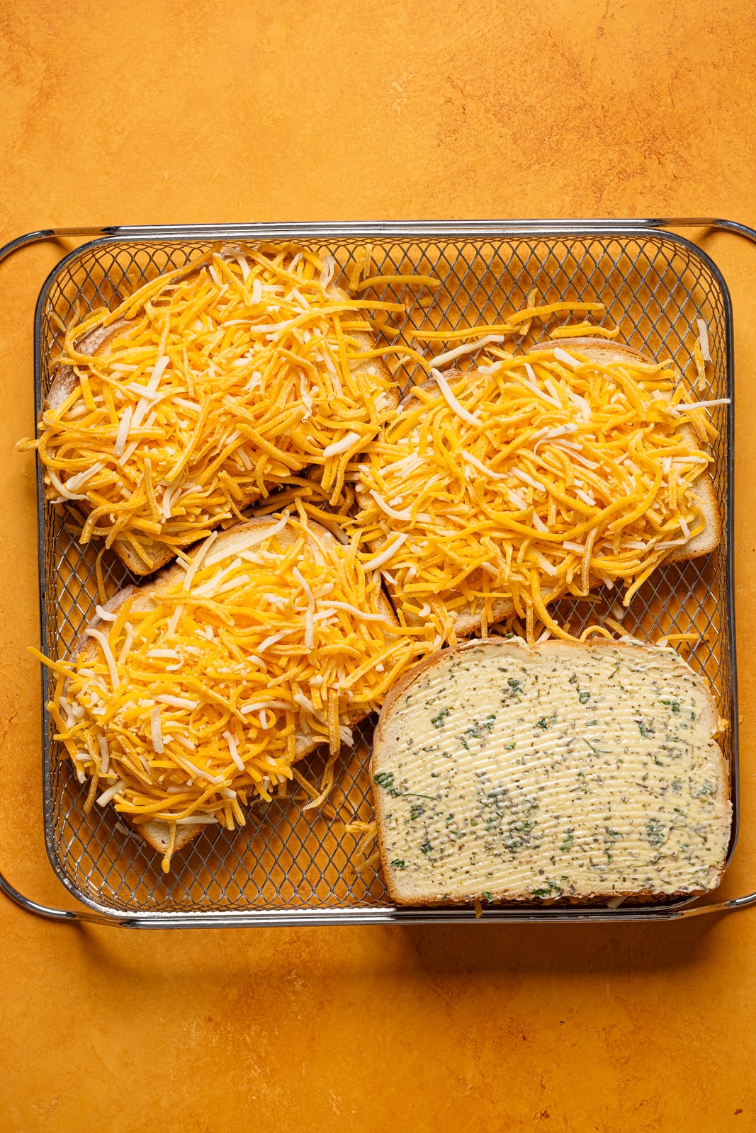 Shredded cheese on slices of bread with herb butter in an Air Fryer basket.