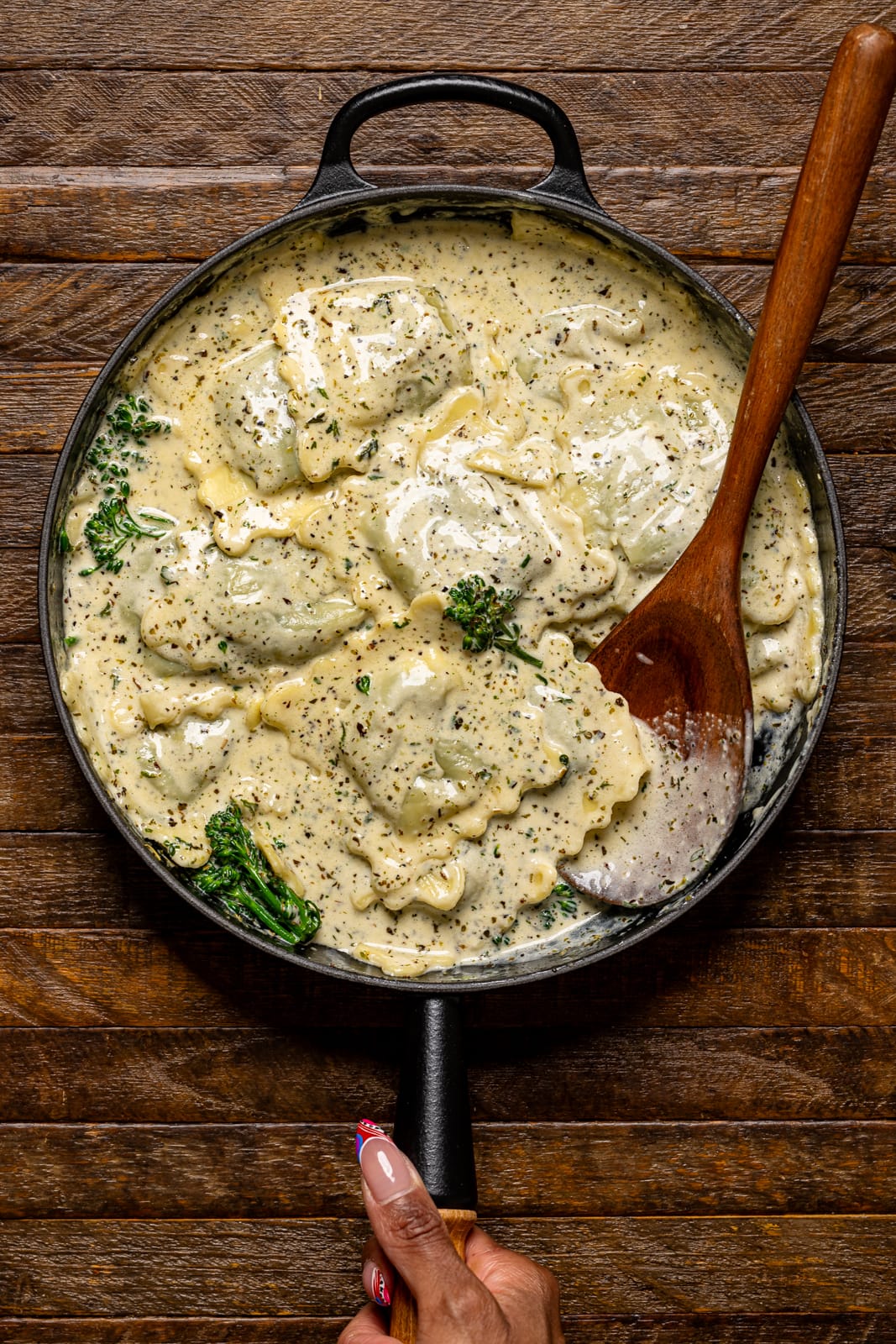 Creamy ravioli in a black skillet being held on a brown wood table.