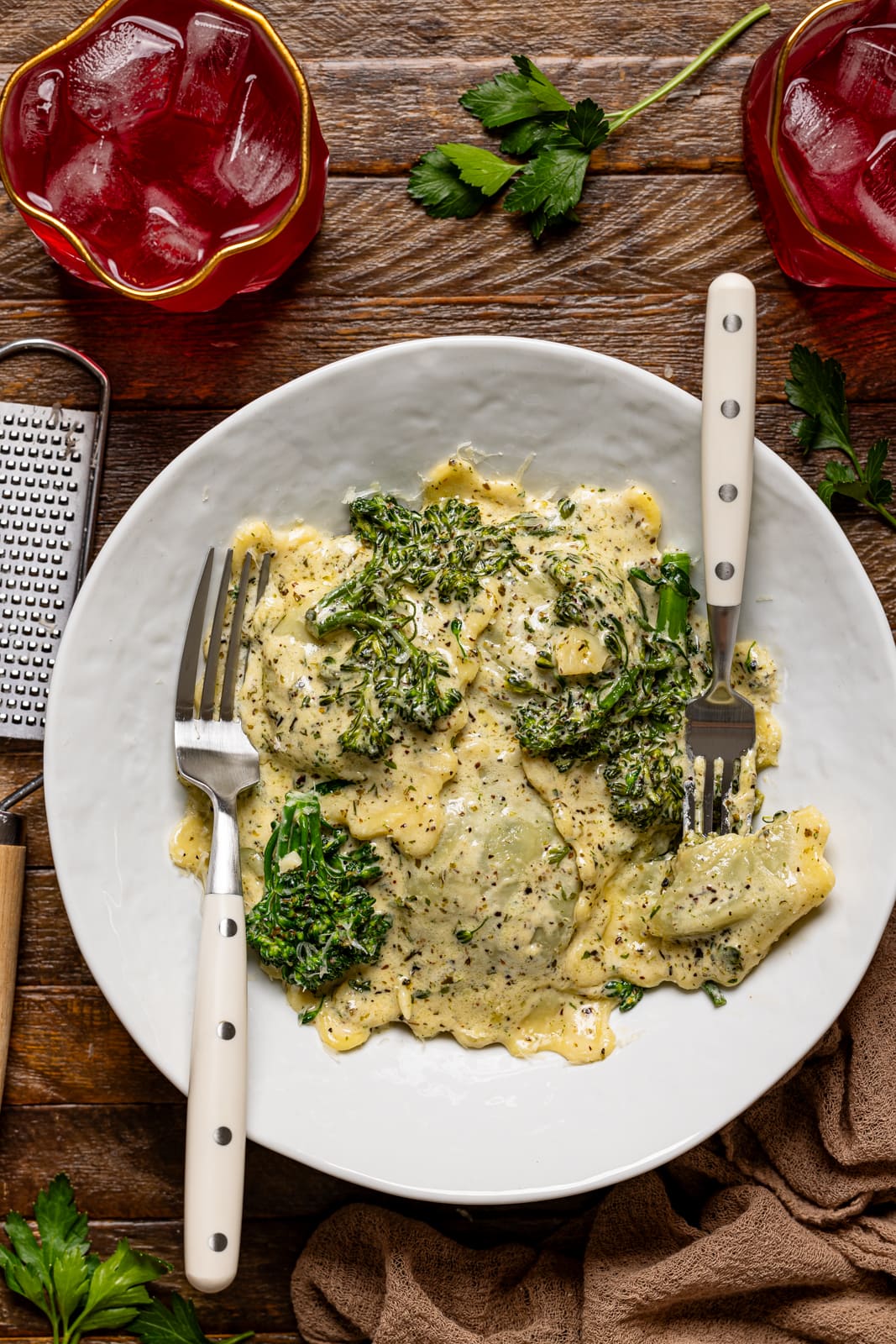 Ravioli in a white plate with two forks, grater, drinks, all on a brown wood table.