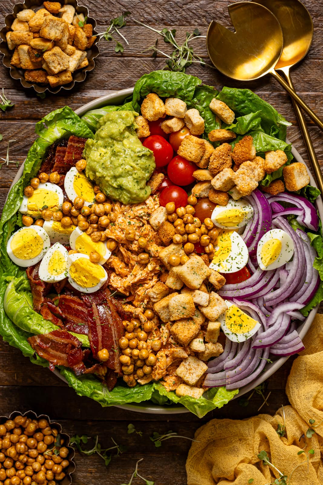 Bowl of cobb salad with gold serving spoons and ingredients.