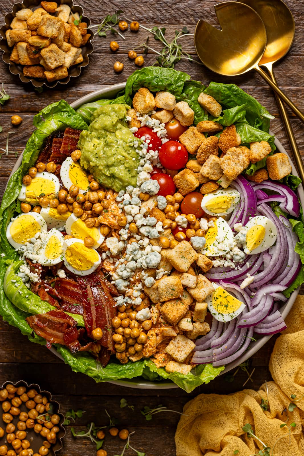 Bowl of cobb salad with spoons, chickpeas and a yellow napkin on a brown wood table.