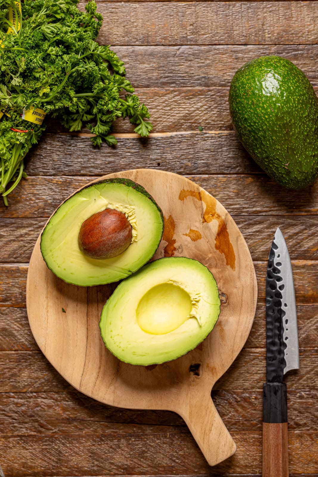 Halved avocados on a cutting board with a knife and ingredients. 